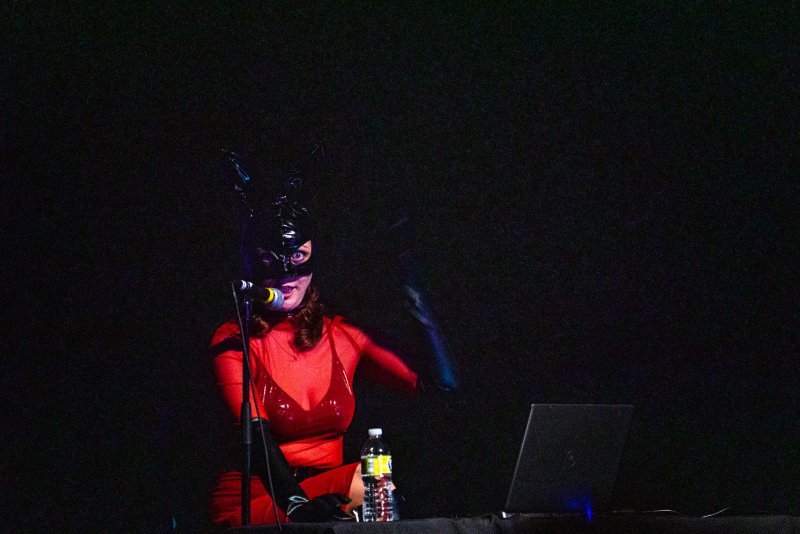 Performer in a shiny black bunny mask and sheer red top, singing into a microphone on a dark stage with a laptop.