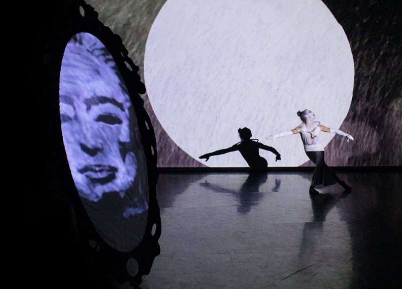 Performance with dancer in white costume, shadow, and mirror reflecting a ghostly face, against a large white circle.