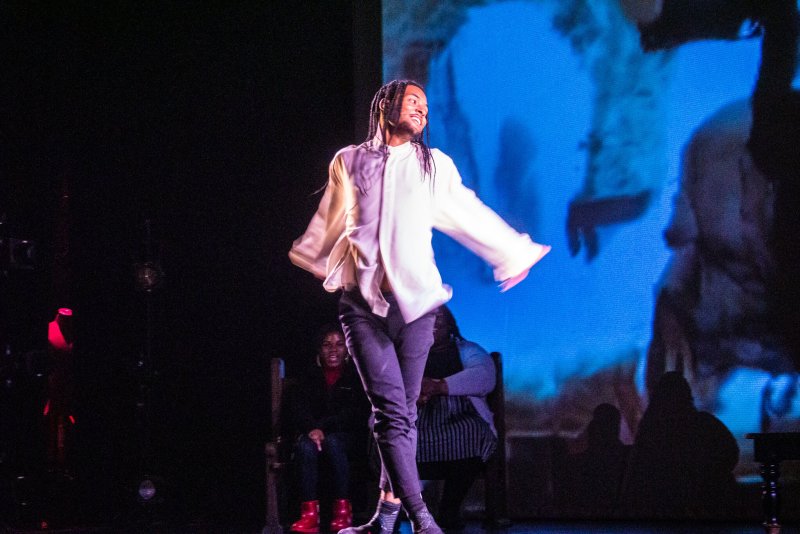Dancer in white shirt and dark pants mid-movement on a dark stage, with a large blue projection screen behind.