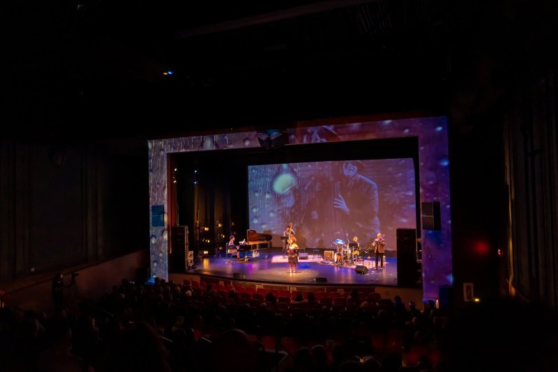 Scott N Andrew's "Suite Life" (2022-2023) shows a band performing on a stage with a large screen displaying a black and white image of a man playing a saxophone, surrounded by purp
