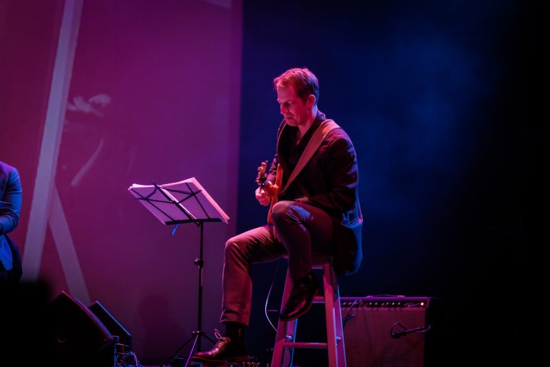 A man in a dark suit sits on a stool, playing an acoustic guitar under purple and blue stage lights, with sheet music on a stand.