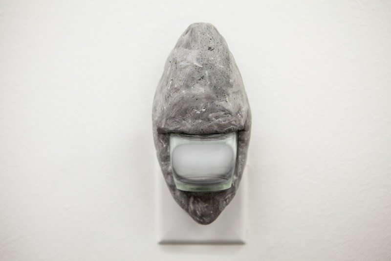 Gray, rock-like sculpture with a clear glass votive holder embedded, containing a white candle, mounted on a white wall.