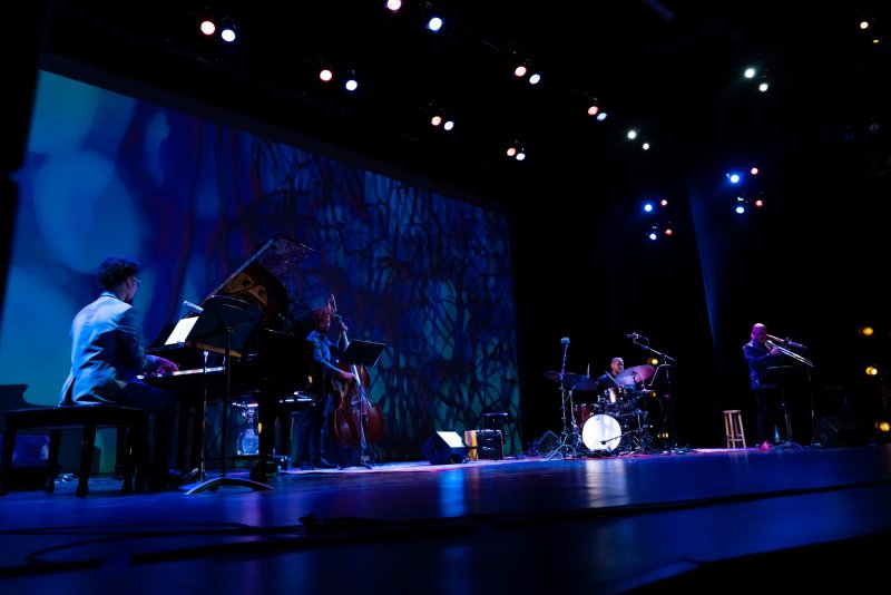 Jazz quartet performs on a dark stage with a large screen displaying abstract blue and purple patterns behind them.
