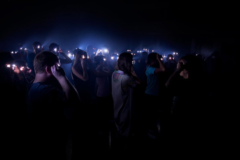 Dark room with a crowd of people holding up phones, their screens illuminating their faces and the space with blue light.