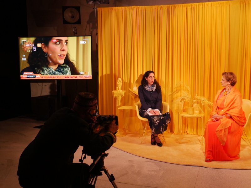Two women in a yellow studio set, one being filmed by a cameraperson, with a live broadcast on a monitor.