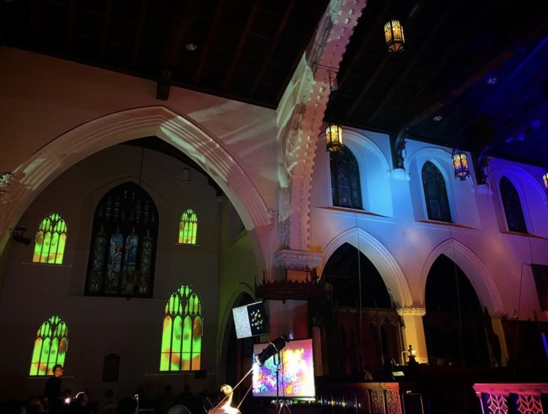 Art installation in a church with colorful projections on arched walls and stained-glass windows, and screens displaying digital art.