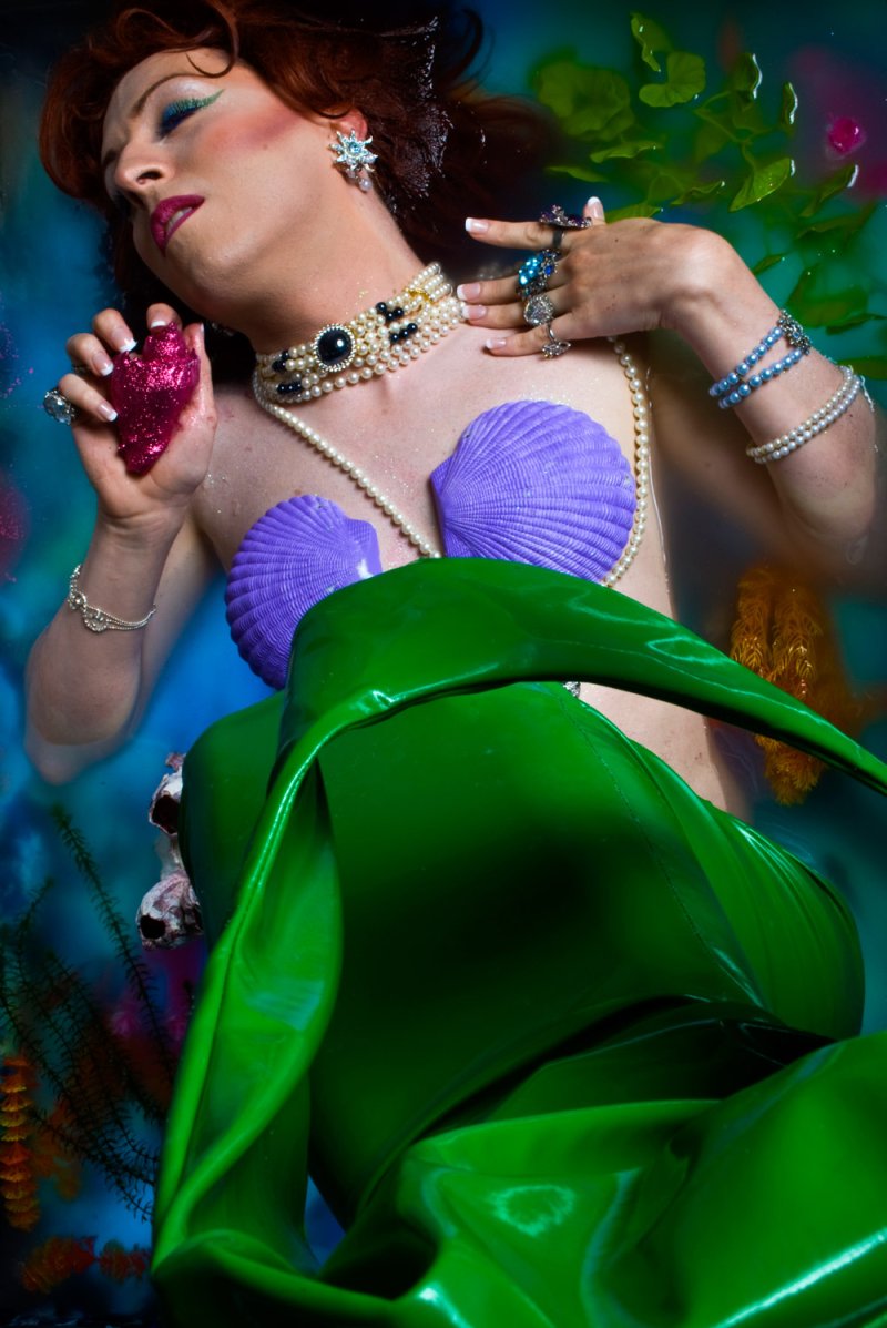 Person in water as mermaid with purple shell bra and green tail, wearing jewelry.