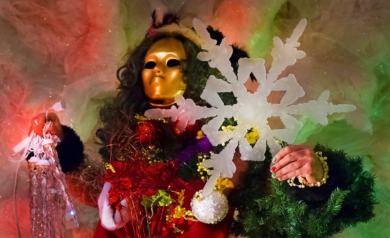 Person in gold mask, dark curly wig, and festive holiday decor holding a large white snowflake and string lights.