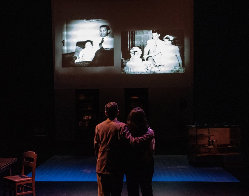 Two people from behind, embracing, watch two black and white photo projections on a dark stage.