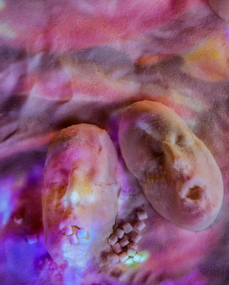 Two sugar-dusted, potato-like forms with facial features, surrounded by sugar cubes on a pink and purple surface.