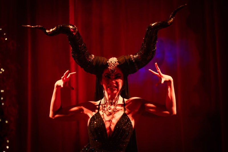 Woman in a black horned headdress, dark makeup, and a sparkly dress, arms raised, against a red curtain background.
