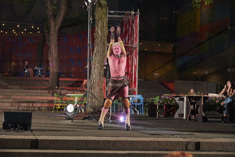 Performance art photograph: A figure with pink skin and blonde hair stands on a stage with arms raised, wearing a brown kilt, knee pads, and heeled boots. The background features trees, seating, and a building with colorful windows.