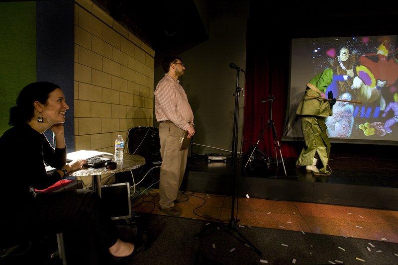 A person in a green costume interacts with a colorful, projected digital art piece on a screen.