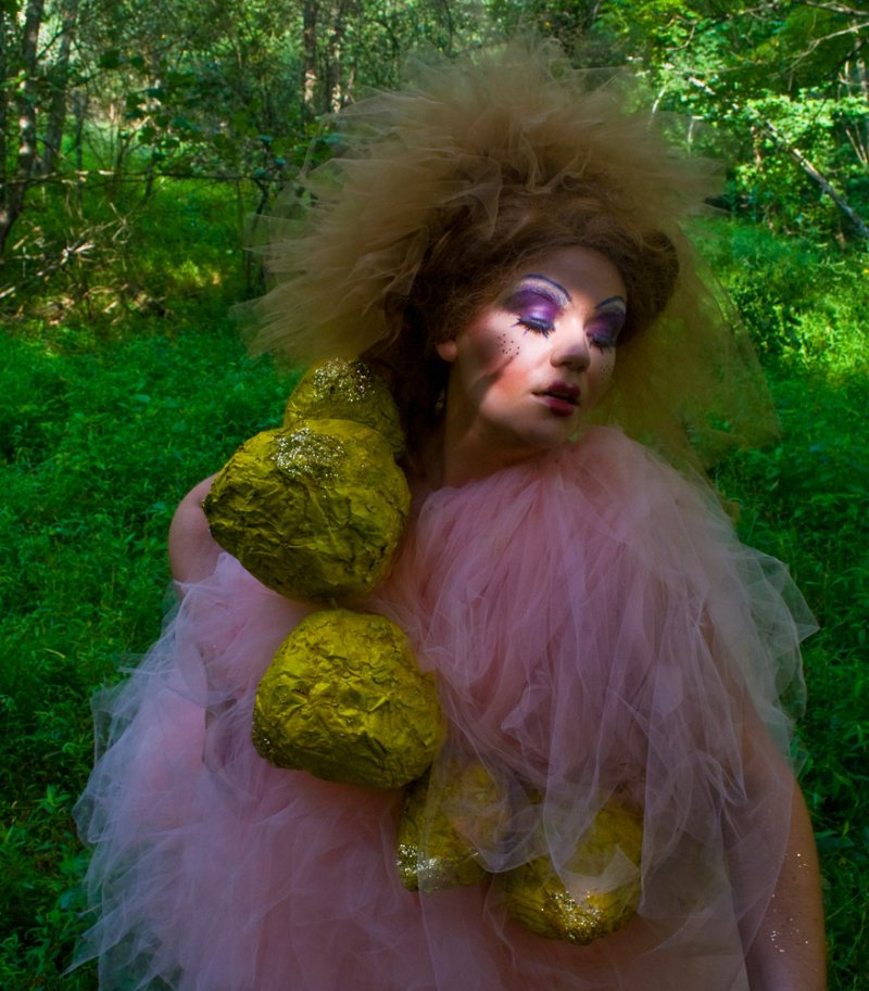 Person in pink tulle dress with large, textured chartreuse forms, dramatic makeup, and voluminous hair in a green forest.