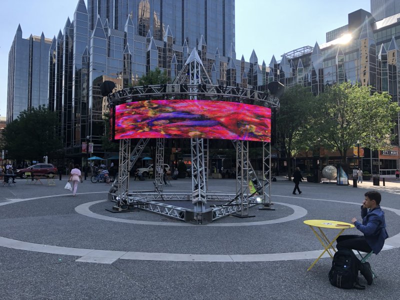 Outdoor video installation: a colorful screen on a metal frame in a plaza with buildings.