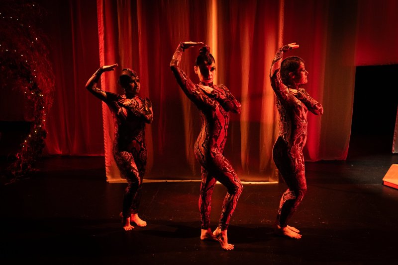 Three dancers in red and black snakeskin bodysuits pose on a dark stage with red lighting and sheer red curtains in the background.