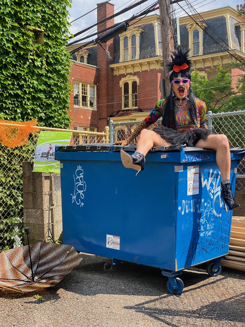 Drag performer in colorful top, black skirt, and heels sits atop a blue dumpster, sticking out their tongue.