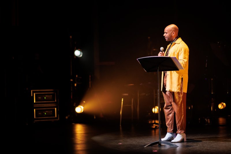 Bald man in yellow jacket and brown pants speaks at a podium on a dark stage, lit by a warm spotlight.
