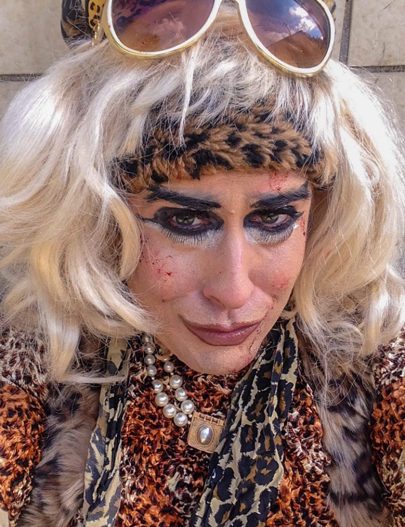 Close-up of a person in drag with blonde hair, leopard print attire, heavy eye makeup, and pearl necklace.