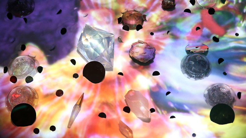 Mixed media installation with various crystals, stones, and glass orbs on a vibrant, colorful projected background.