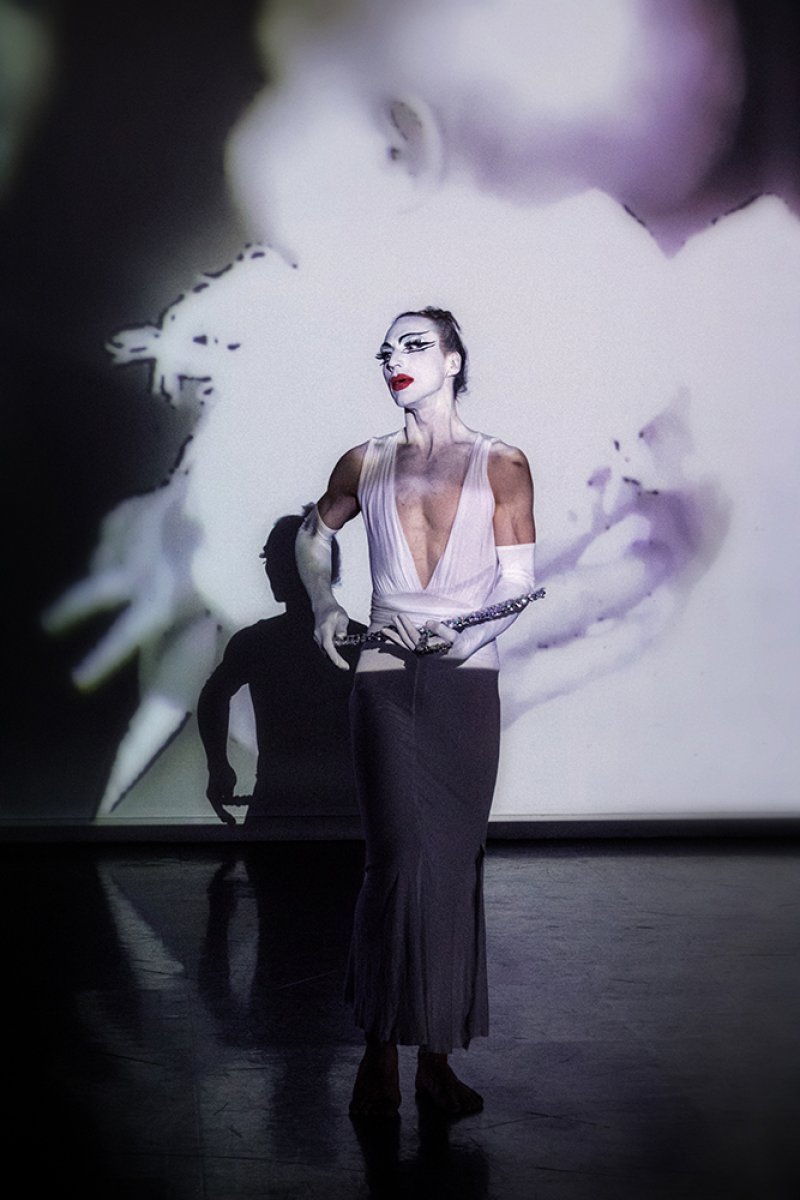 Performer in white makeup, deep v-neck top, and long skirt, holding a flute, against a projected abstract background.