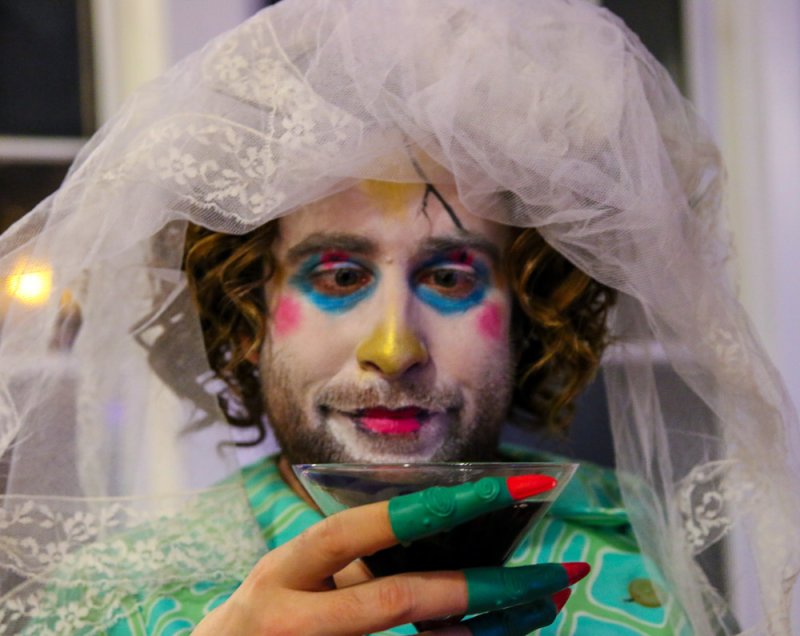 A person in a white veil and theatrical makeup, holding a martini glass with green-painted fingers.