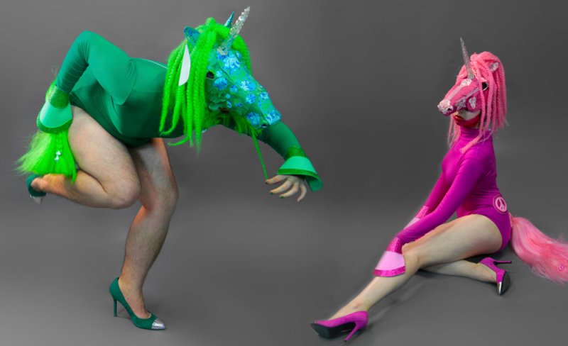 Two people in unicorn costumes, one green with a blue floral head, the other pink with a peace sign.