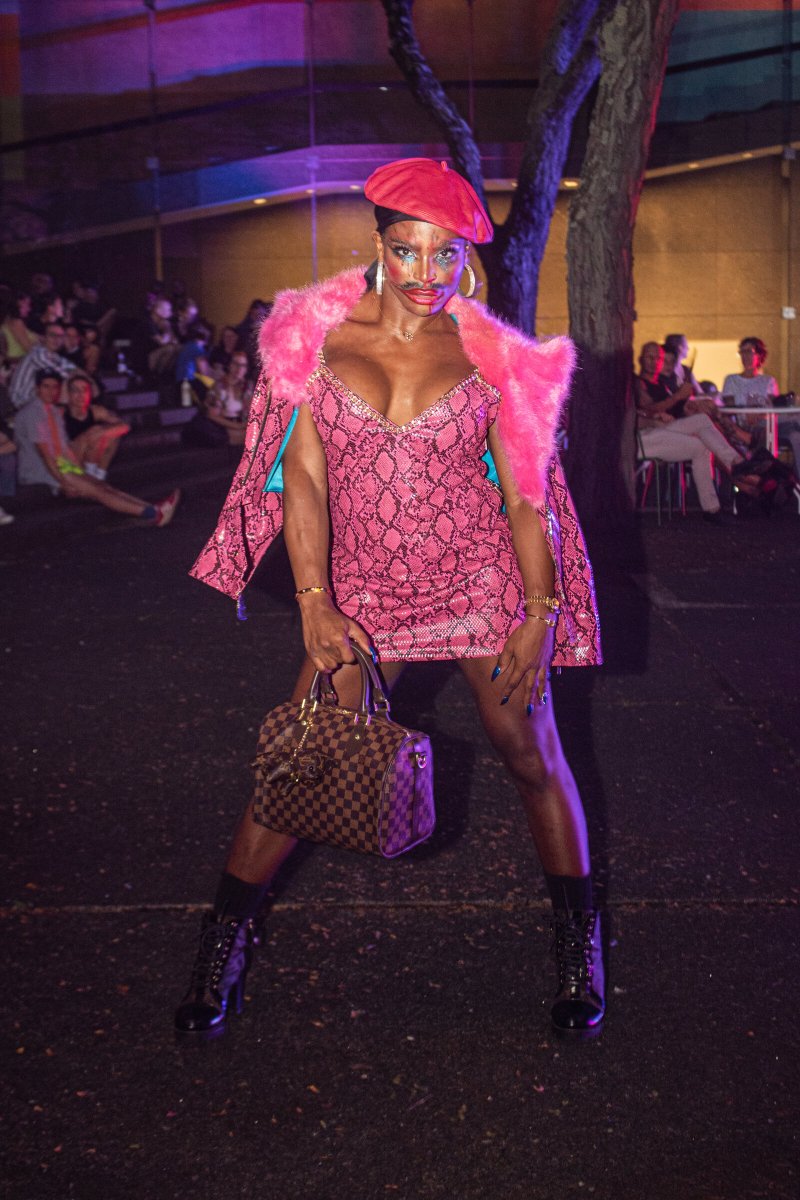Drag performer in a pink snakeskin dress, pink fur stole, red beret, and combat boots, holding a handbag.