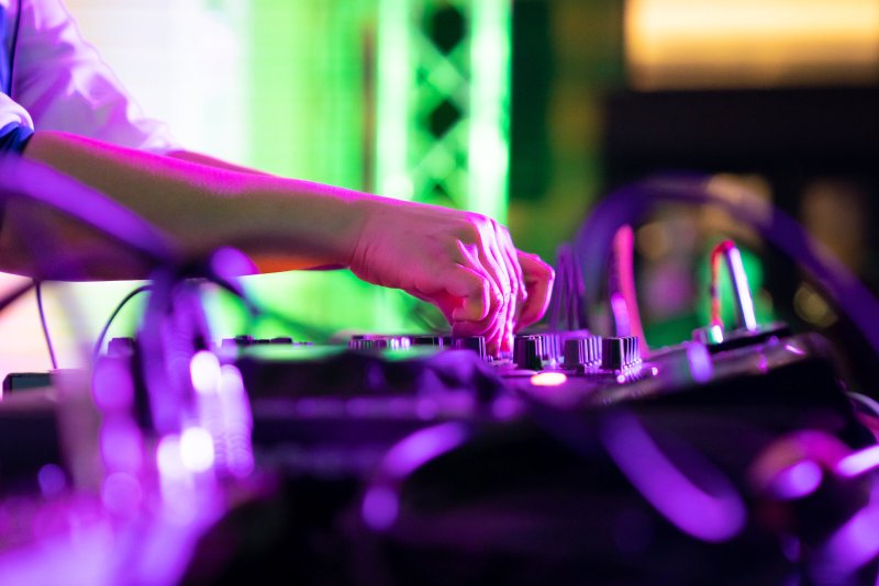Close-up of hands manipulating a DJ mixer under vibrant pink and green lighting. The mixer features numerous knobs and buttons, with blurred cables and background elements.