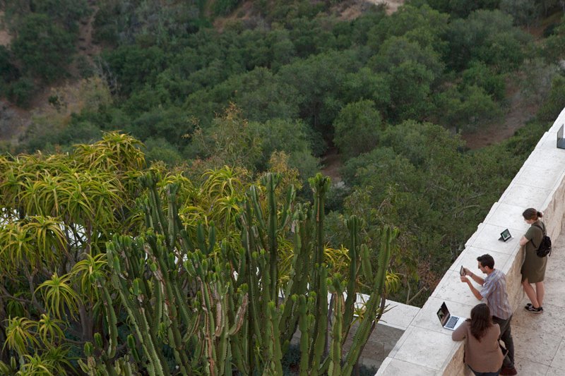 Overhead view of three people on a balcony overlooking a lush green forest, interacting with laptops and phones.