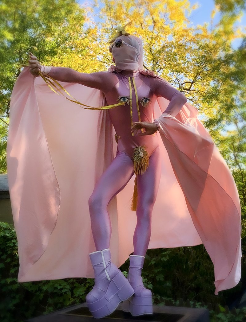 Full shot of a figure in a pink bodysuit and cape, wearing platform boots, against a green and yellow tree backdrop. The figure has a veiled head, sunglasses, and decorative eyes and tassels on the suit.