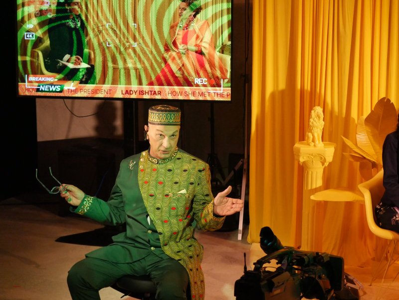 Performance artist in a green and gold patterned suit and hat, gesturing, with a news broadcast screen behind them.