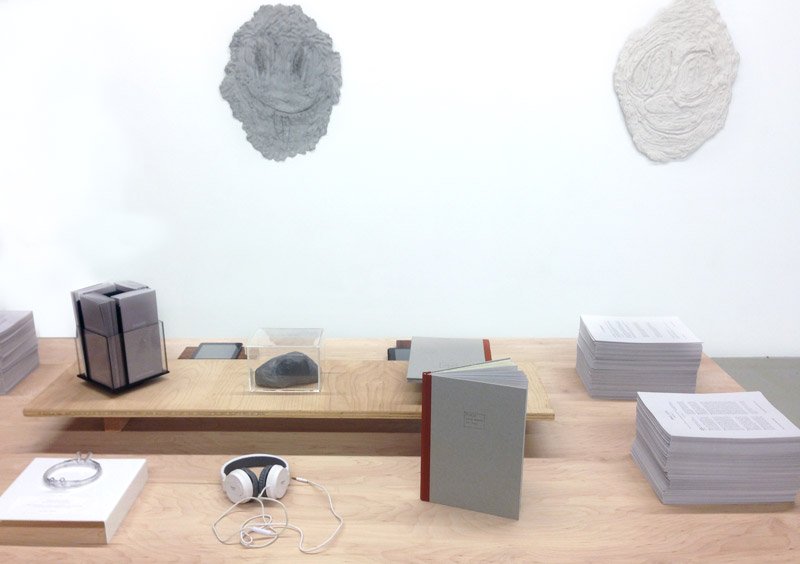 Installation with a light wood table displaying books, papers, headphones, and a rock, beneath two textured gray wall hangings.