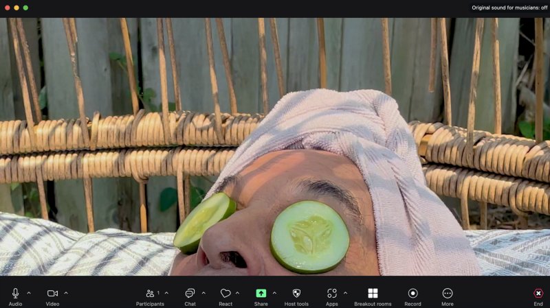Close-up of a person with cucumber slices on their eyes, a towel on their head, in a wicker chair, viewed through a video call interface.