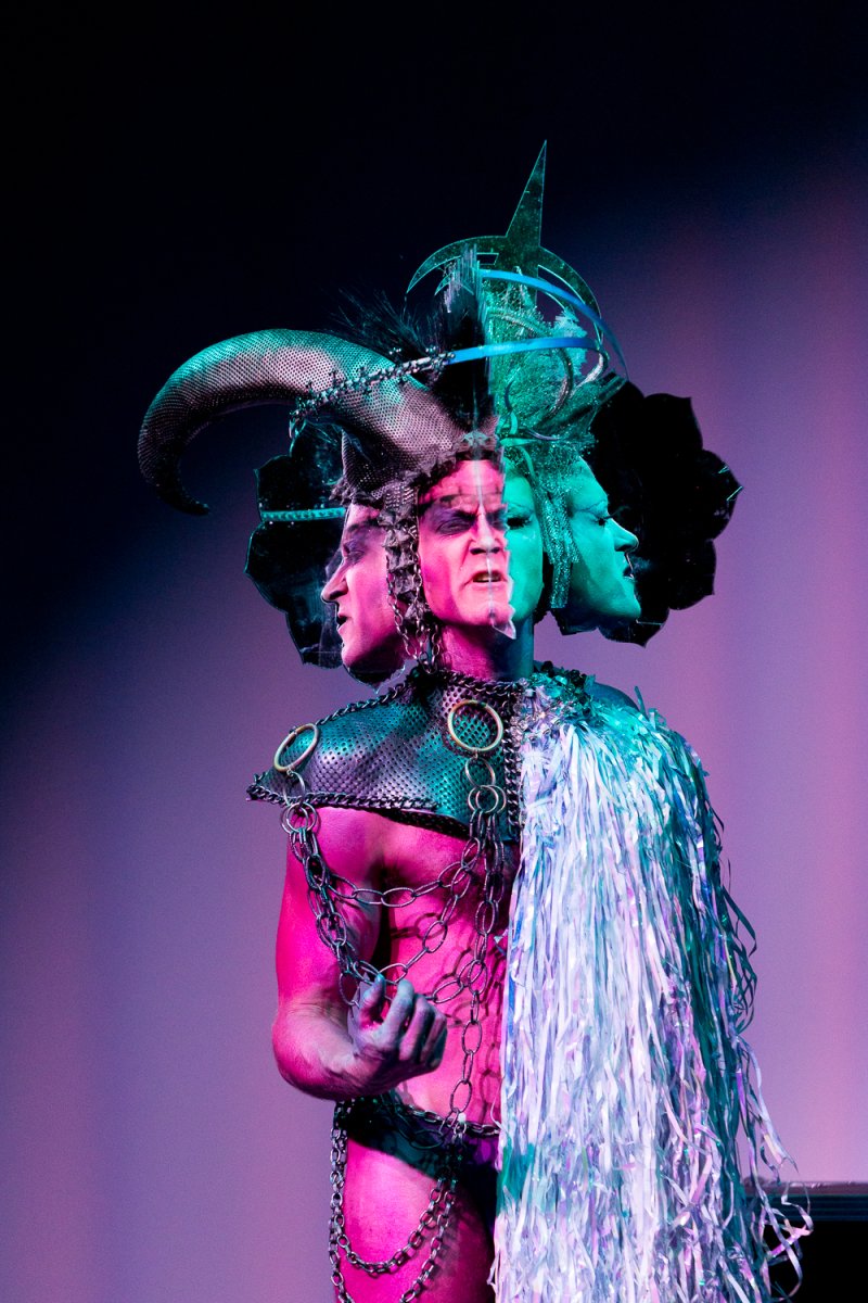 Drag performer Darrell Thorne in elaborate costume with multiple faces, horns, chains, and tinsel, lit with pink and blue light.