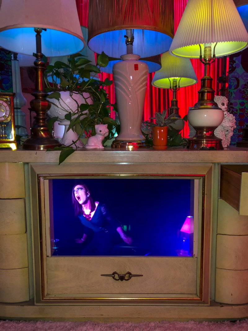 Installation with a vintage dresser displaying a video of a person singing in blue light. Lamps, plants, and figurines adorn the dresser top, creating a cluttered, colorful aesthetic.