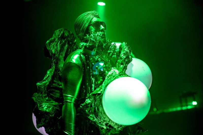 Performer in green metallic costume with large white spheres, dark sunglasses, and a green spotlight.