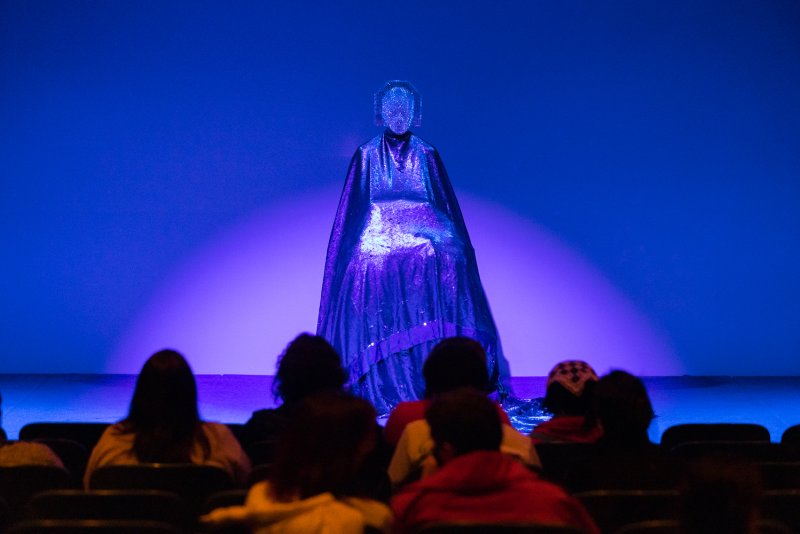 Performer in a shimmering blue-purple costume and mask, seated on a stage bathed in blue light, facing an audience.