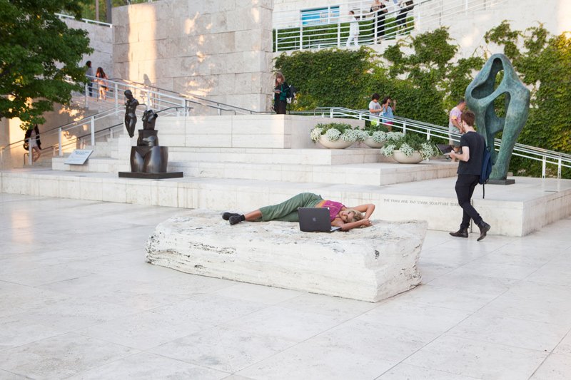 A person lies on a large stone block with a laptop, surrounded by modern sculptures and people on stairs in a sunny outdoor plaza.