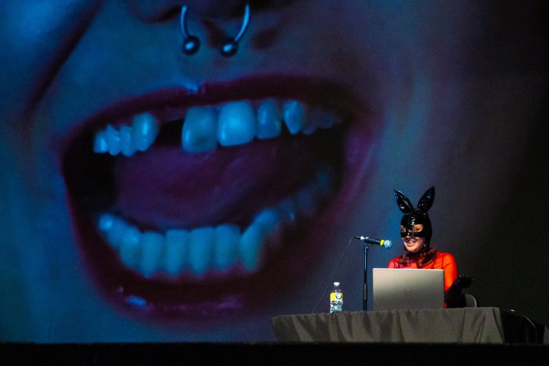 Installation featuring a person in a bunny mask at a table with a laptop, set against a large projected image of an open mouth with a nose ring, lit in blue and purple.