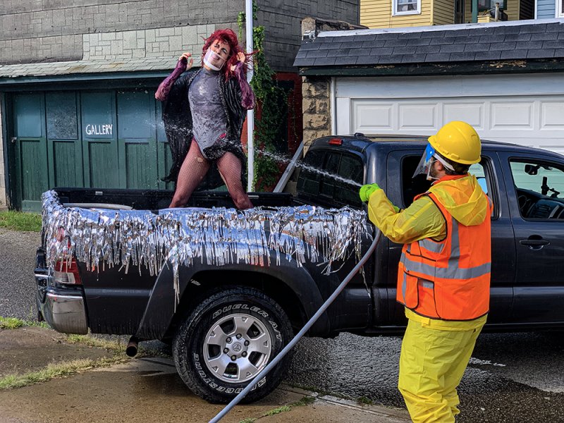 Outdoor scene: a person in work gear hoses down a figure in a sequined outfit and fishnets standing in a pickup truck bed decorated with silver fringe. A "Gallery" sign is visible on a building in the background.
