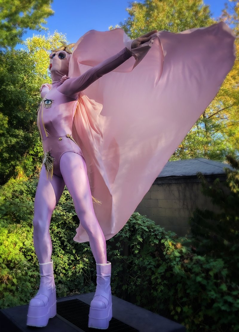 Person in a light pink bodysuit and platform boots stands outdoors with arms outstretched, holding a flowing pink cape. The figure has a covered face with sunglasses and decorative eye and feather embellishments.