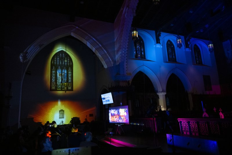 Dark church interior with colorful light projections on walls, stained glass, and two screens displaying abstract digital art.