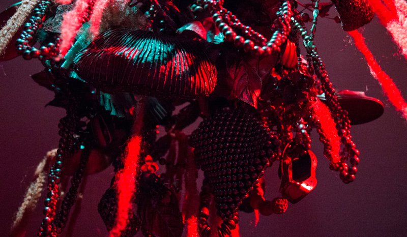 Close-up of a dark, textured sculpture with hanging beaded strands, illuminated by strong red and teal lighting.