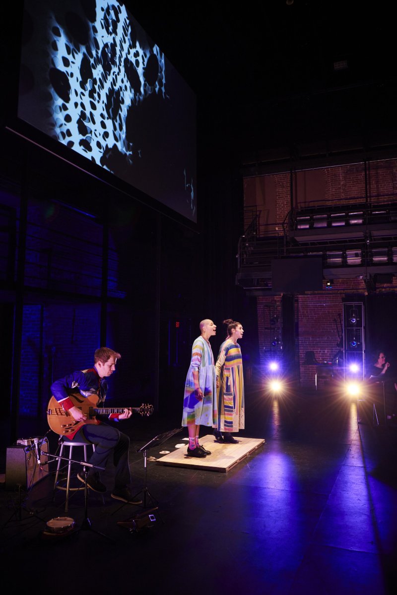 Performance scene with guitarist, two singers in colorful striped dresses on a small platform, and a screen showing blue and white abstract patterns. Stage lit with blue hues and spotlights. Brick wall visible in the background.