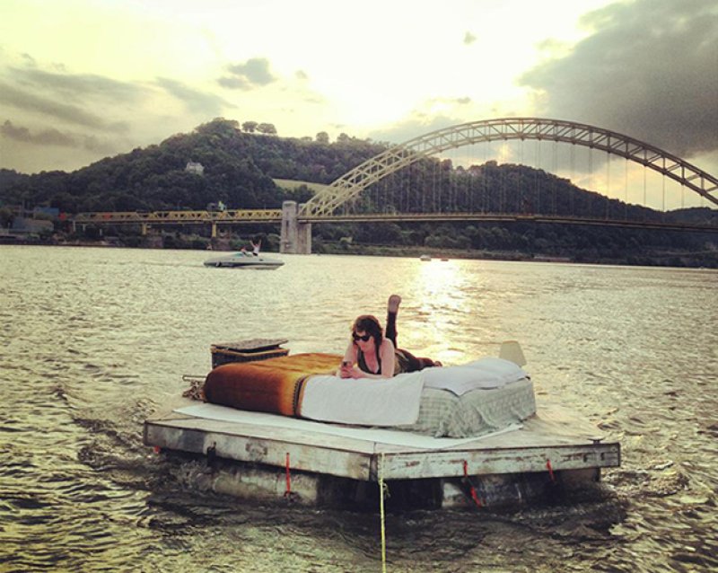 Woman in sunglasses on a bed on a raft, floating on a river with a bridge and hills in the background at sunset.