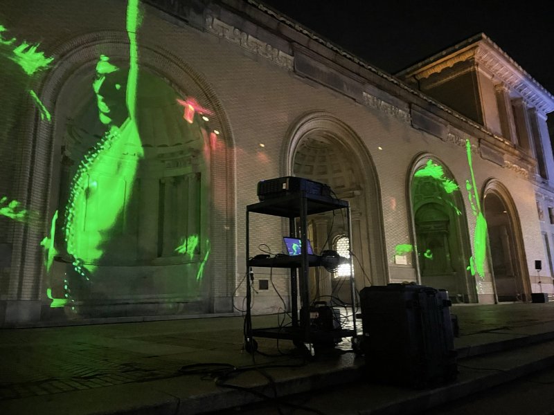 Green digital projections of abstract figures on a brick building at night, with a projector setup in the foreground.