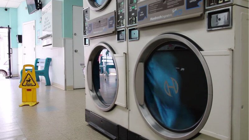 Laundromat interior with two large washing machines in the foreground, a yellow wet floor sign, and a blue bench in the background.