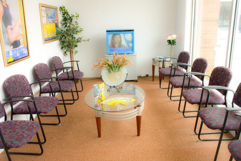 A waiting room with patterned chairs, a glass coffee table with magazines, and a TV displaying a woman.