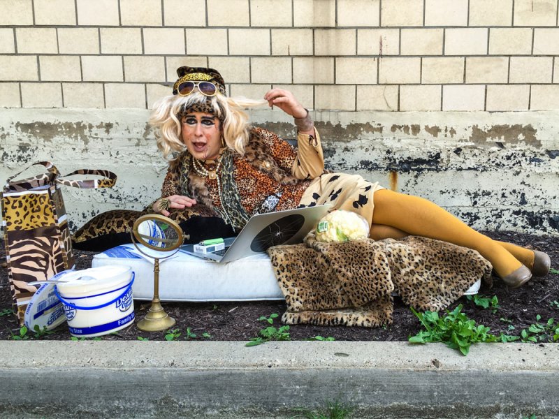 Drag queen in leopard print, blonde wig, and sunglasses reclines on a mattress outdoors, surrounded by props.